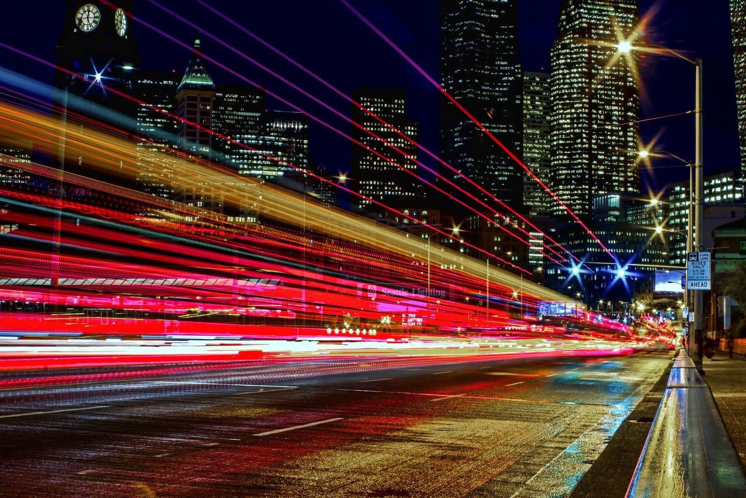 Long-exposure traffic light trails streaking along a wet city street below a lit-up skyline at night