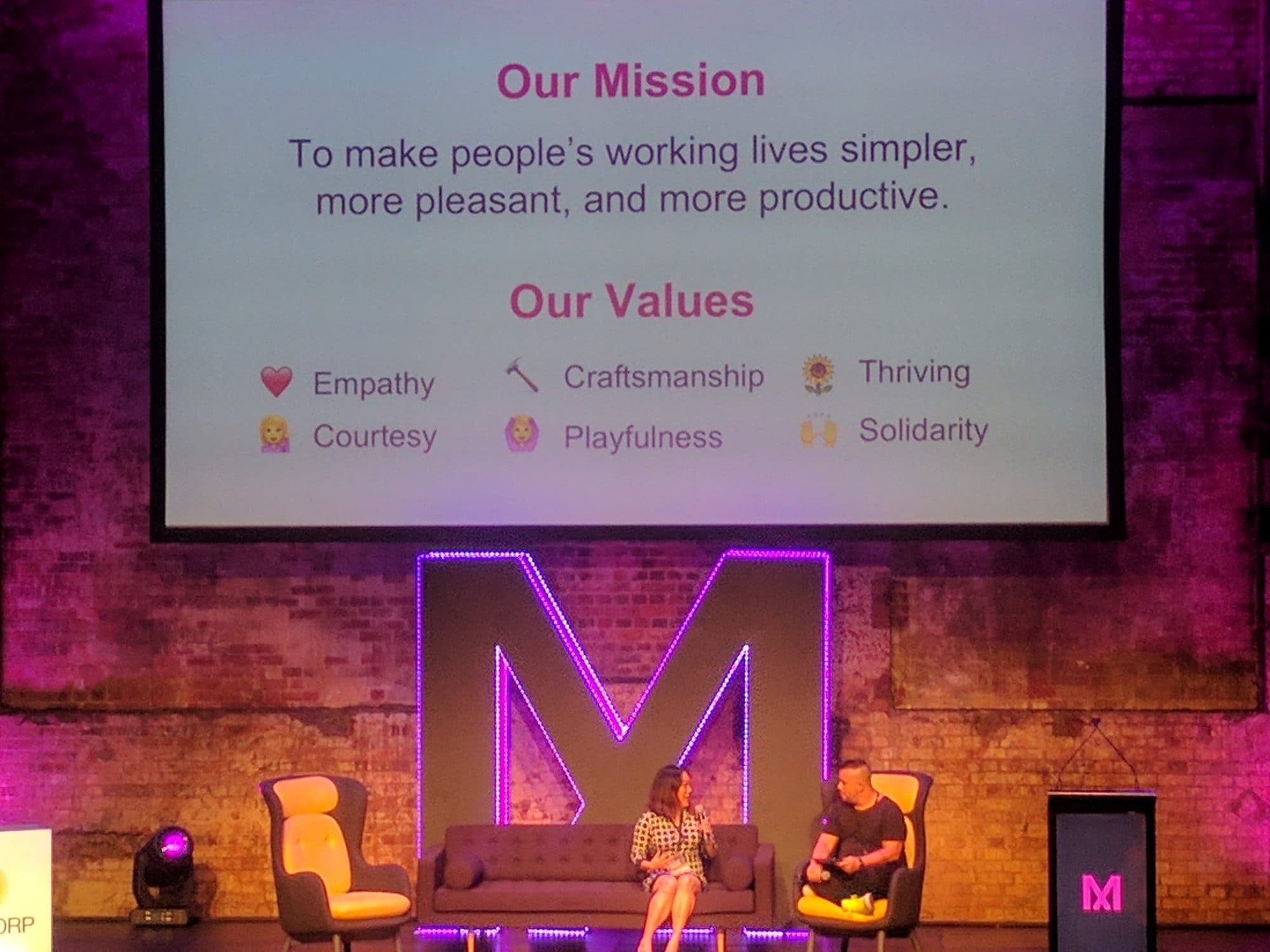 Two speakers in a stage interview at Myriad beneath a slide reading Our Mission and Our Values, lit M logo behind them