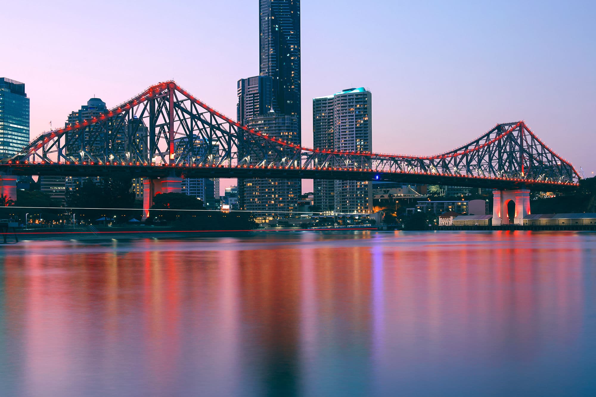 The Story Bridge in Brisbane at dusk, lit red, with the city skyline behind it and the river in front.