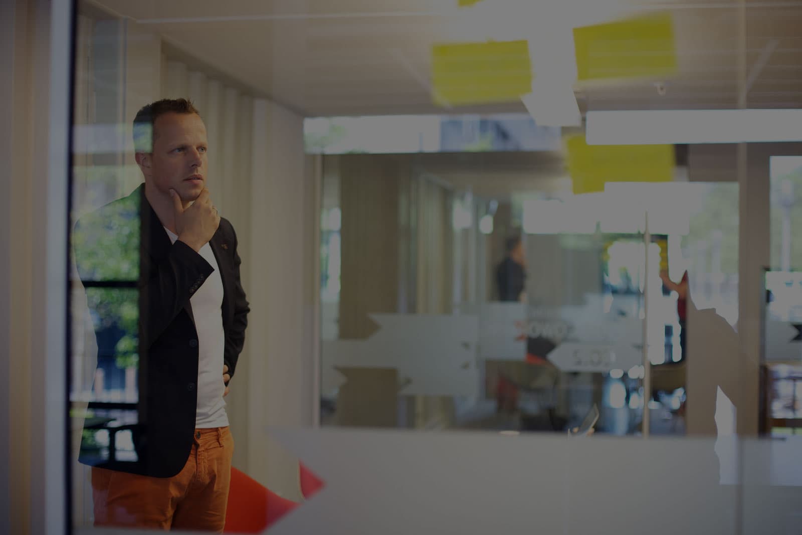 A man in a dark blazer stands behind a glass office wall covered in yellow sticky notes, hand on his chin, looking past the camera into an open-plan office.