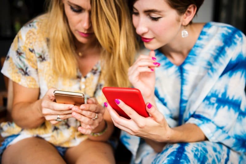 Two young women sitting side by side, each looking down at her smartphone