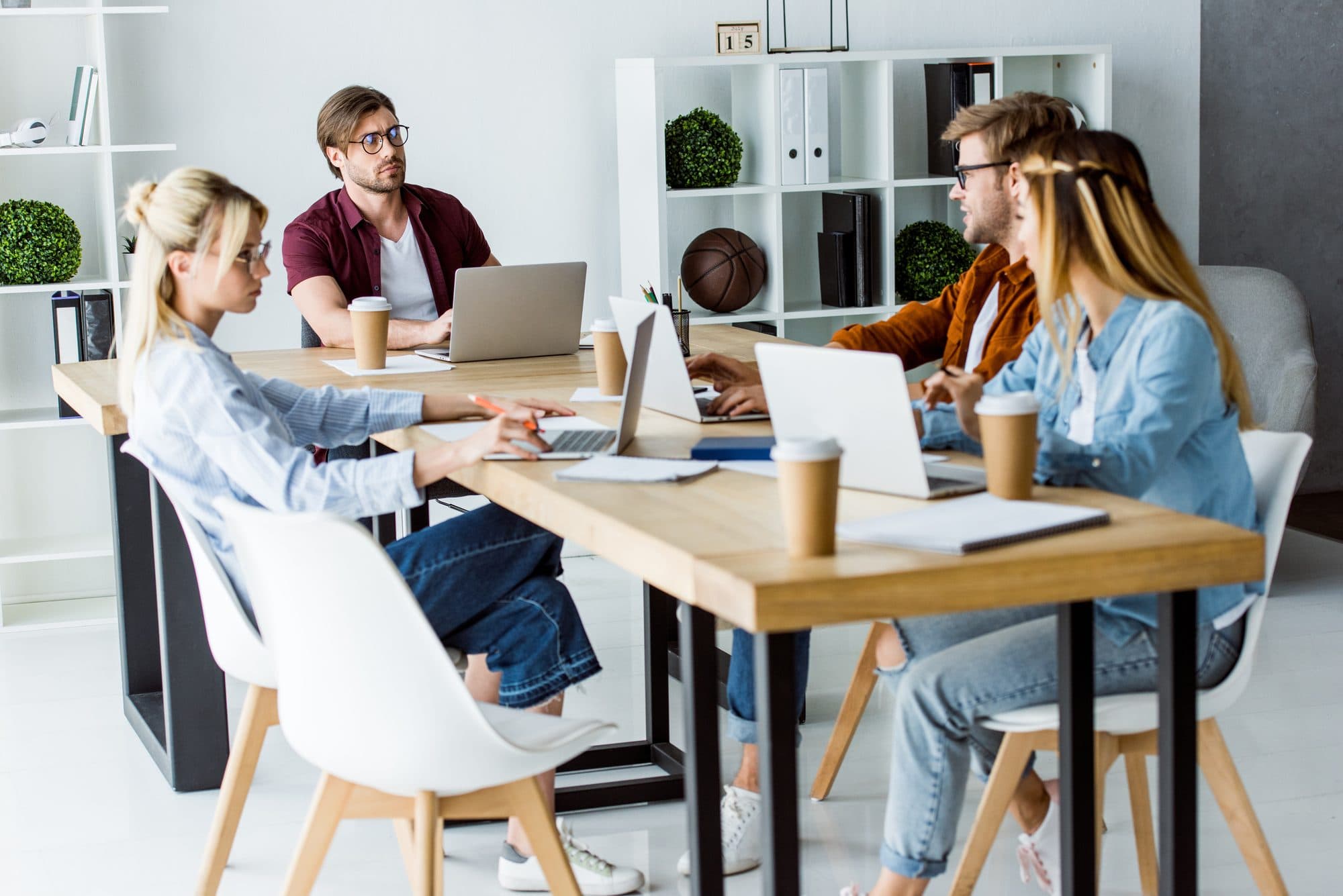 Five people talking around a boardroom table with open laptops, notepads and takeaway coffee cups
