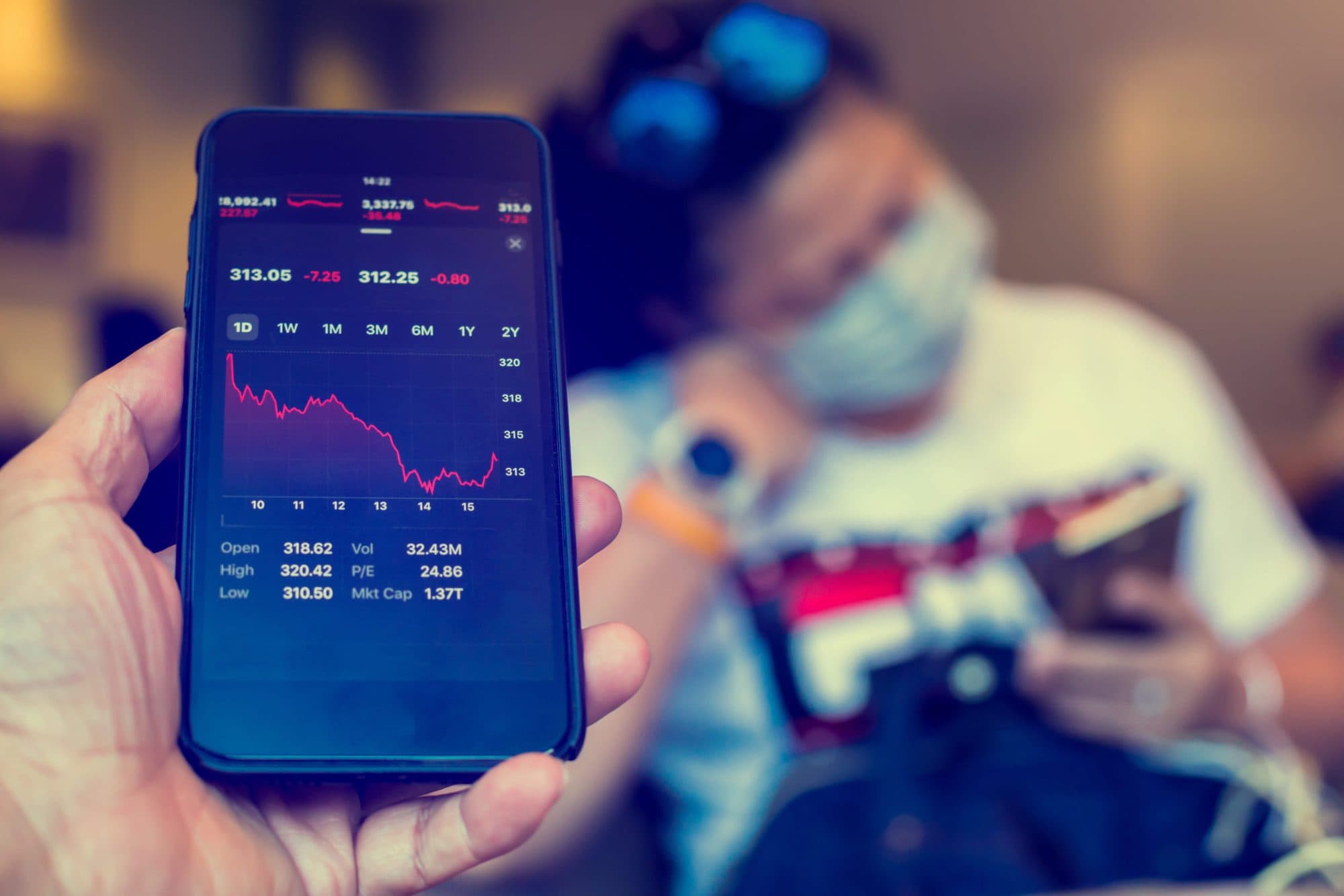 Hand holding a phone showing a falling stock chart in red, with a masked man on his phone blurred in the background