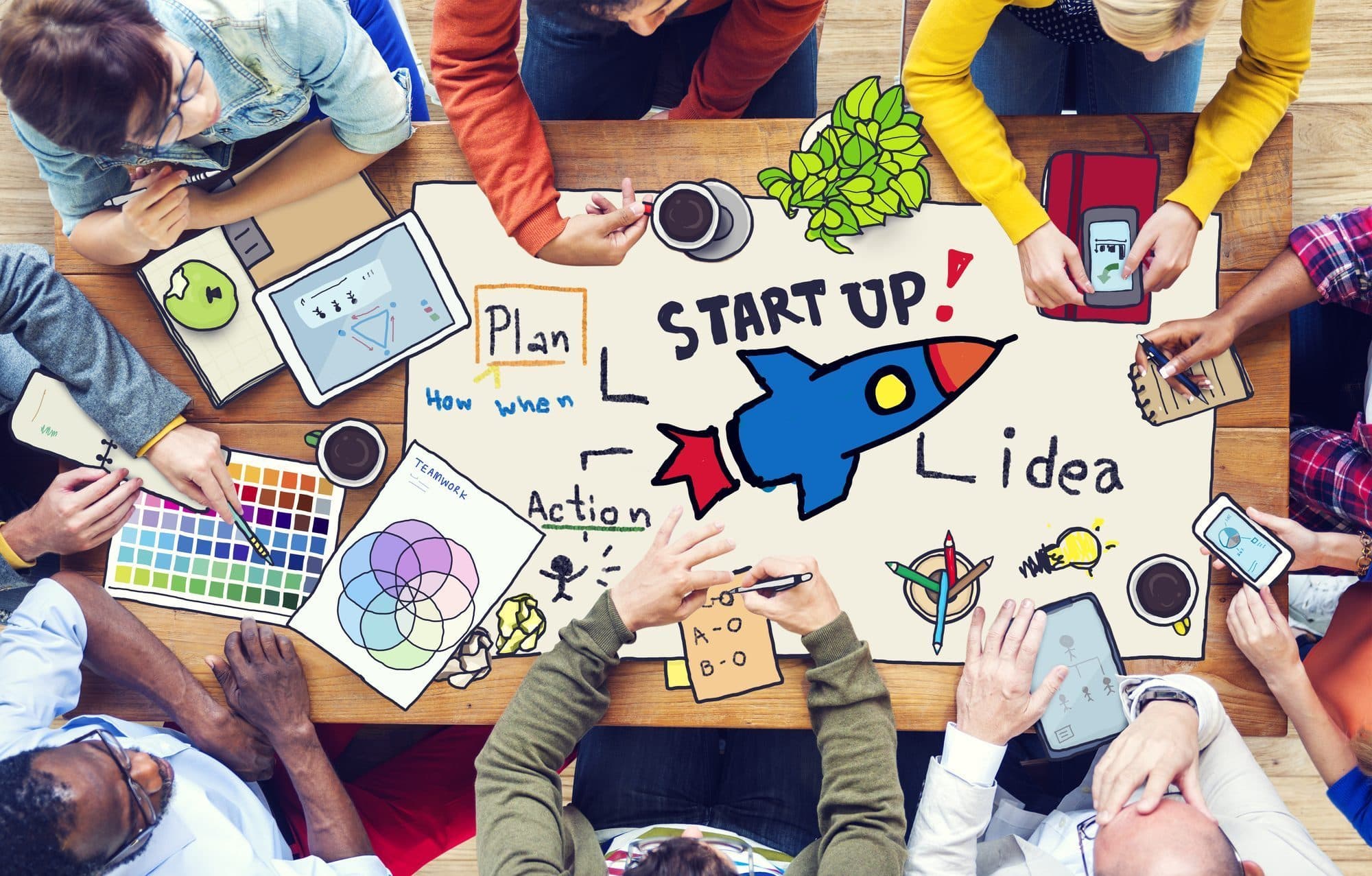 Overhead view of a team around a table covered with hand-drawn startup doodles of a rocket, plan, action and idea notes