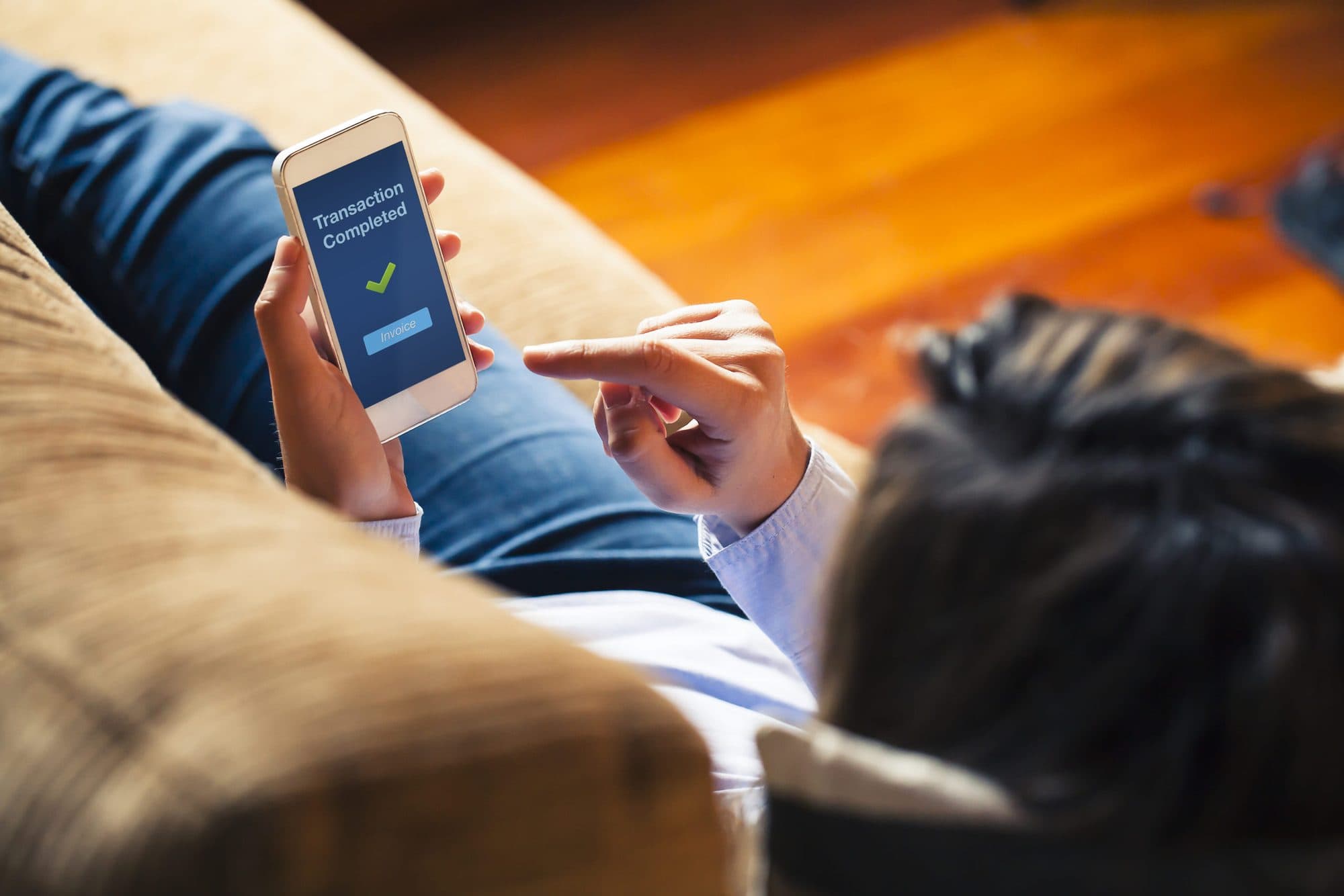 Person on a sofa tapping a phone that displays a Transaction Completed message with a green tick and an invoice button
