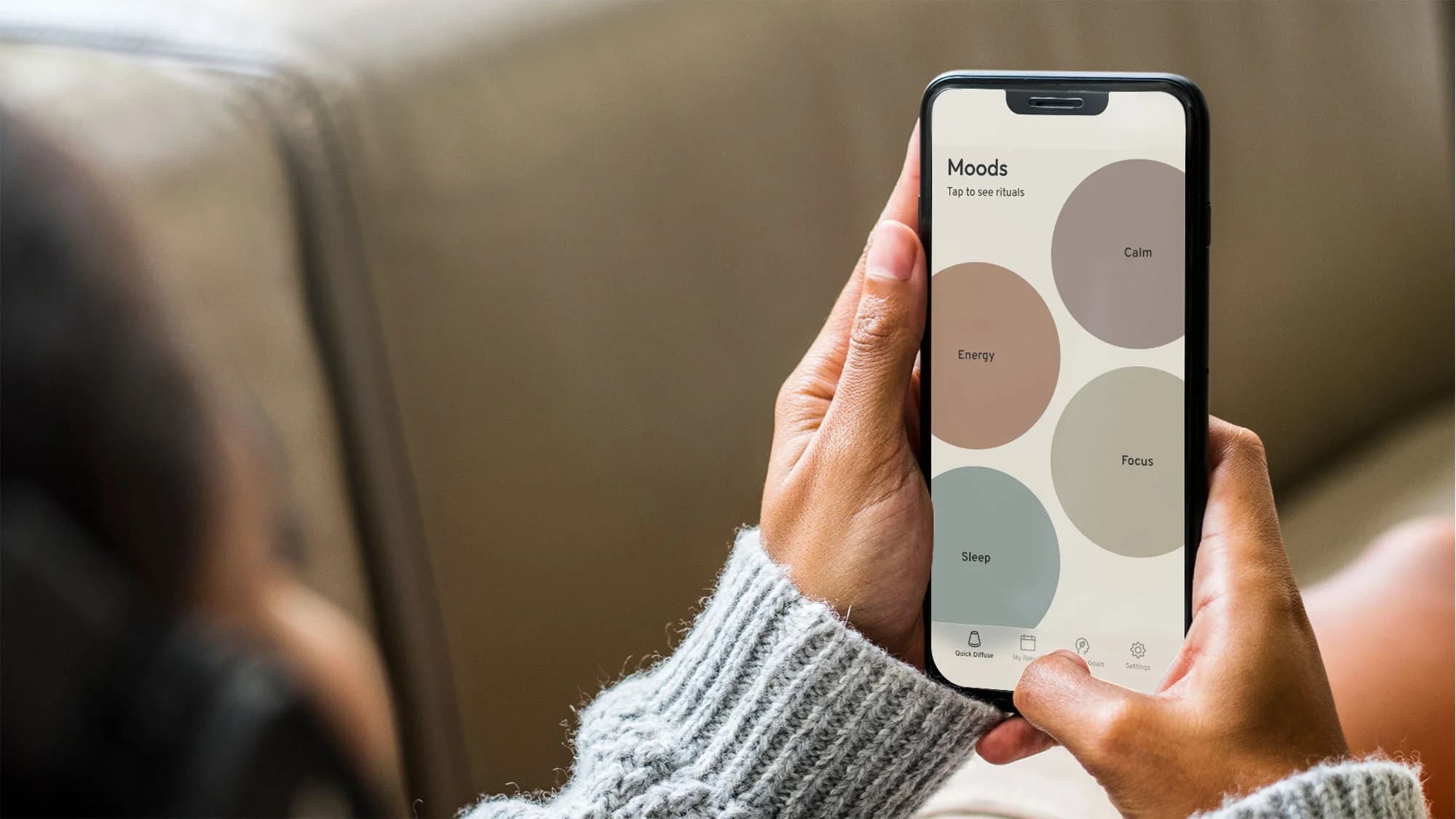 Woman on a sofa holding a phone showing the Auric app Moods screen with Calm, Energy, Focus and Sleep circles to tap for rituals